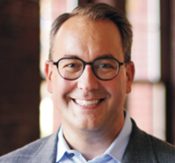 Head-and-shoulders portrait of a smiling man with glasses in a blazer and light blue shirt.