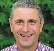Smiling man with short gray hair wearing a checkered shirt outdoors, green foliage in background