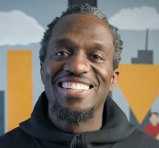Smiling man facing the camera outdoors, wearing a black hoodie, with a colorful city backdrop behind him.
