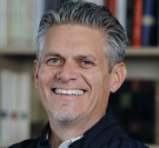Portrait of a smiling man with gray hair and a light goatee, wearing a dark jacket, with a library backdrop.