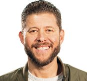 Close-up of a smiling man with short dark hair and a beard, wearing a light jacket, against a white background.