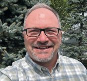 Portrait of a smiling man wearing black-rimmed glasses and a plaid shirt, outdoors with evergreen trees in the background.