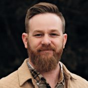 Smiling man with light brown hair and a full beard, wearing a beige jacket over a plaid shirt, outdoors.